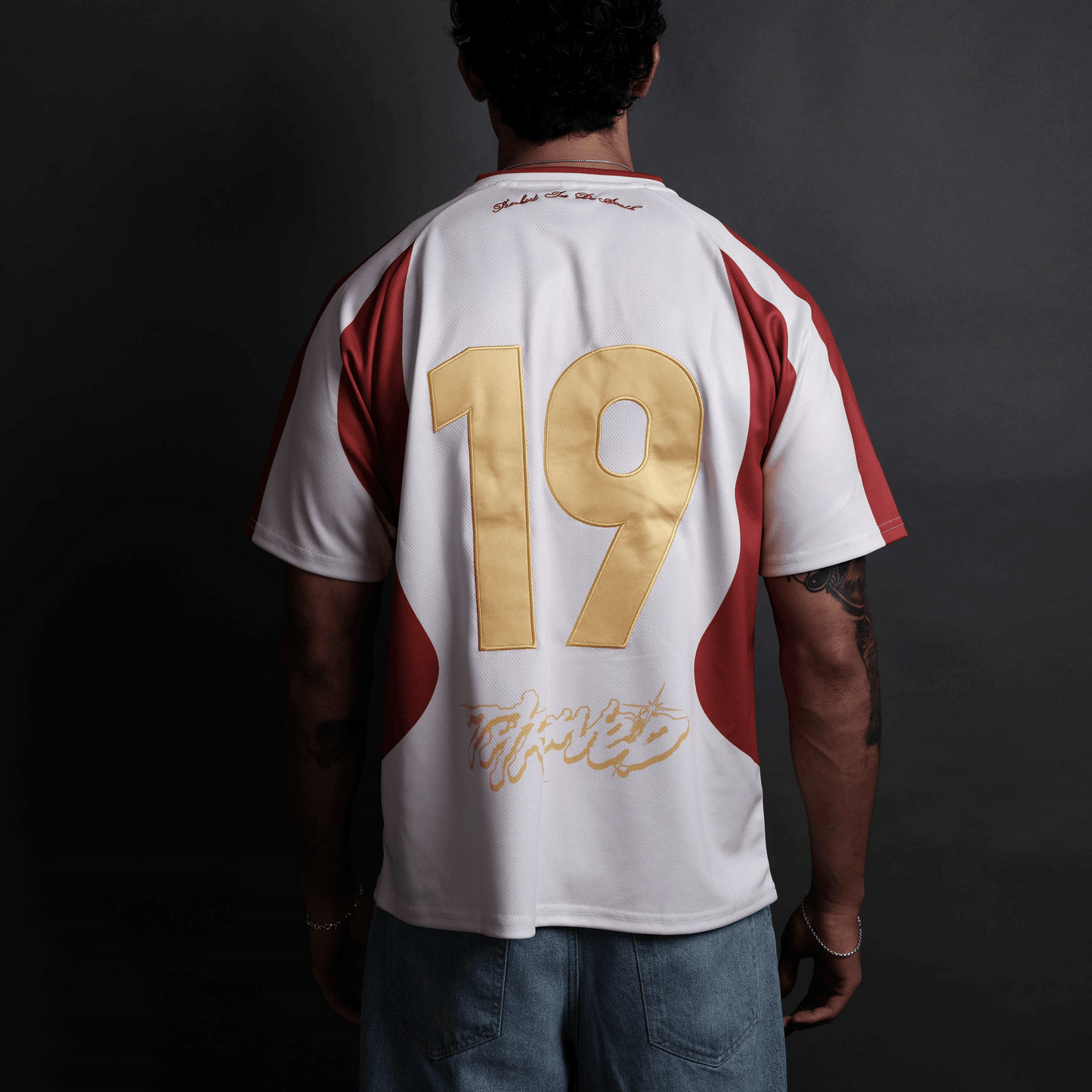 A man viewed from behind wearing a white short-sleeve jersey with red side panels and a large metallic gold number 19 on the back, paired with blue jeans against a dark studio background.