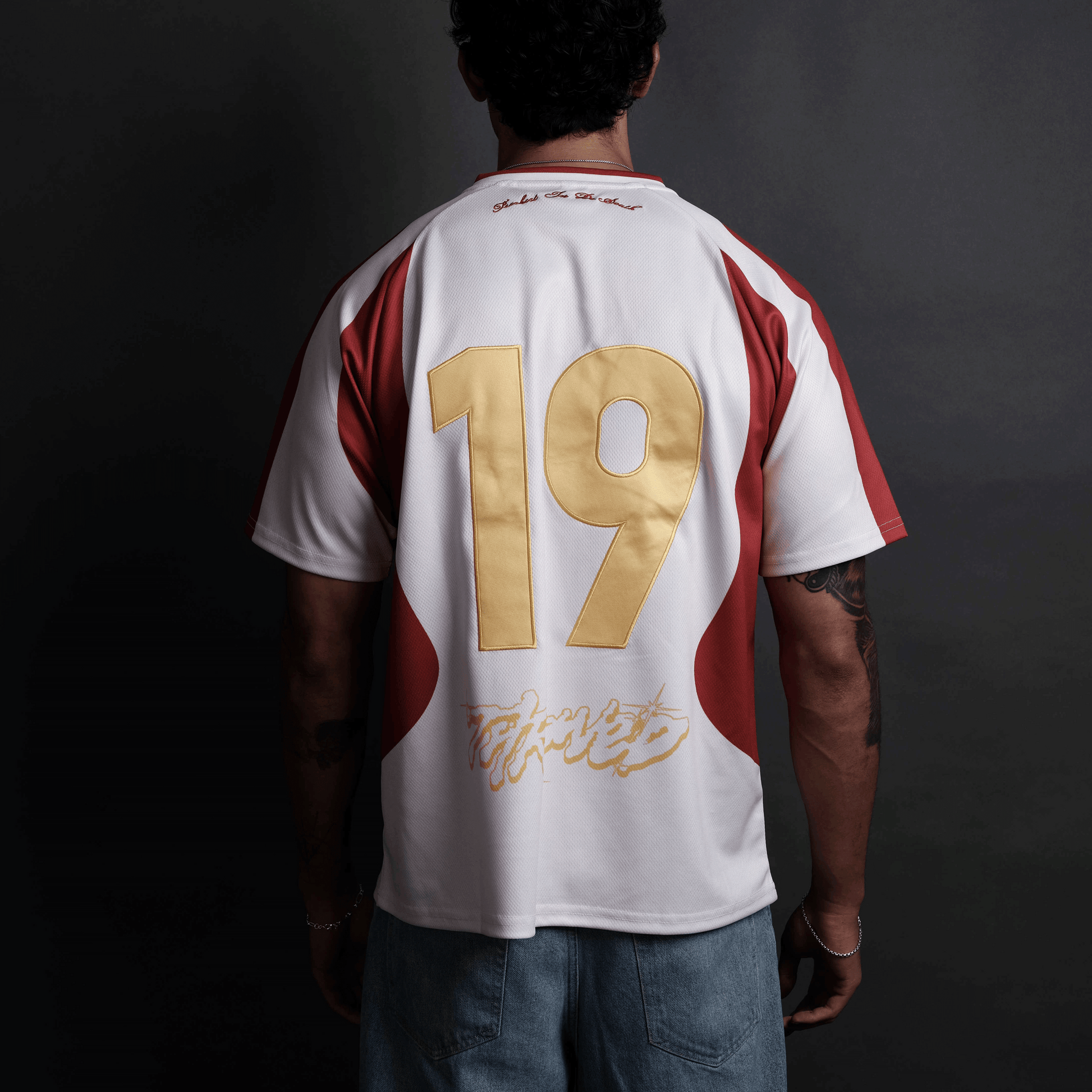 A man viewed from behind wearing a white short-sleeve jersey with red side panels and a large metallic gold number 19 on the back, paired with blue jeans against a dark studio background.